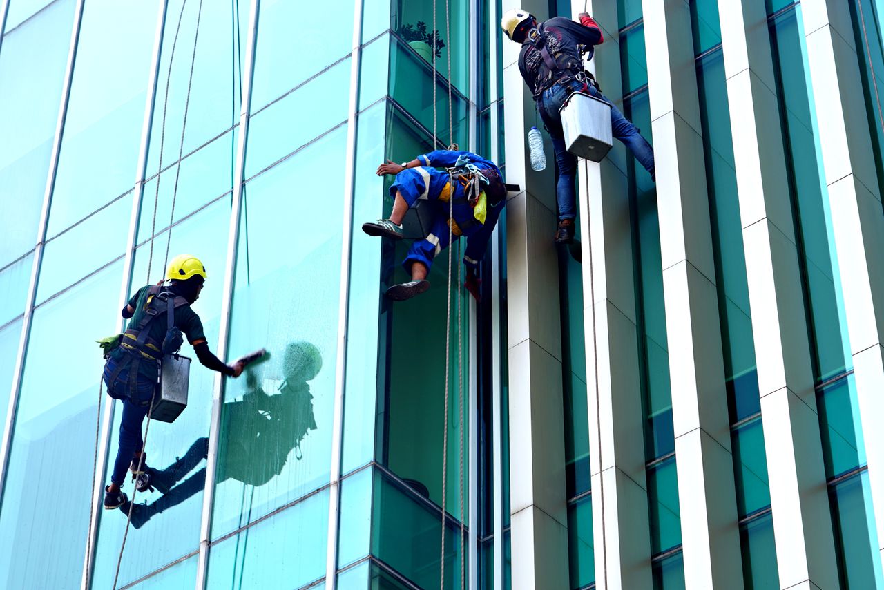Trois laveurs de vitres suspendus à des cordes nettoient, en journée, la façade d’un immeuble moderne en verre.